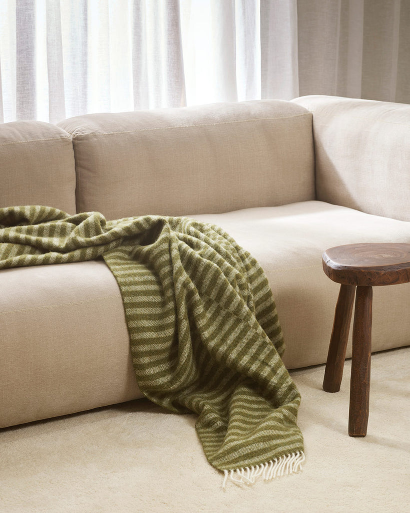 An olive green throw blanket with striping in a lighter green and cream twisted edging in 100% wool draped over a cream coloured couch. 