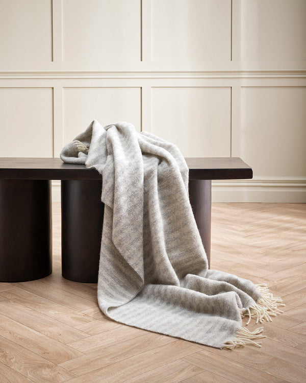 A soft 100% wool throw blanket draped over a designer chair, with silver striping and cream twisted edging. 