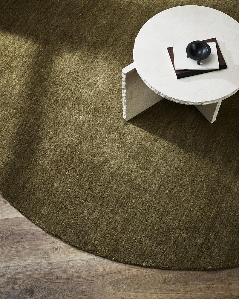 A designer coffee table seen from above. Under it is a gorgeous round warm green toned 100% wool floor rug in Moss by Baya