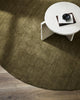 A designer coffee table seen from above. Under it is a gorgeous round warm green toned 100% wool floor rug in Moss by Baya