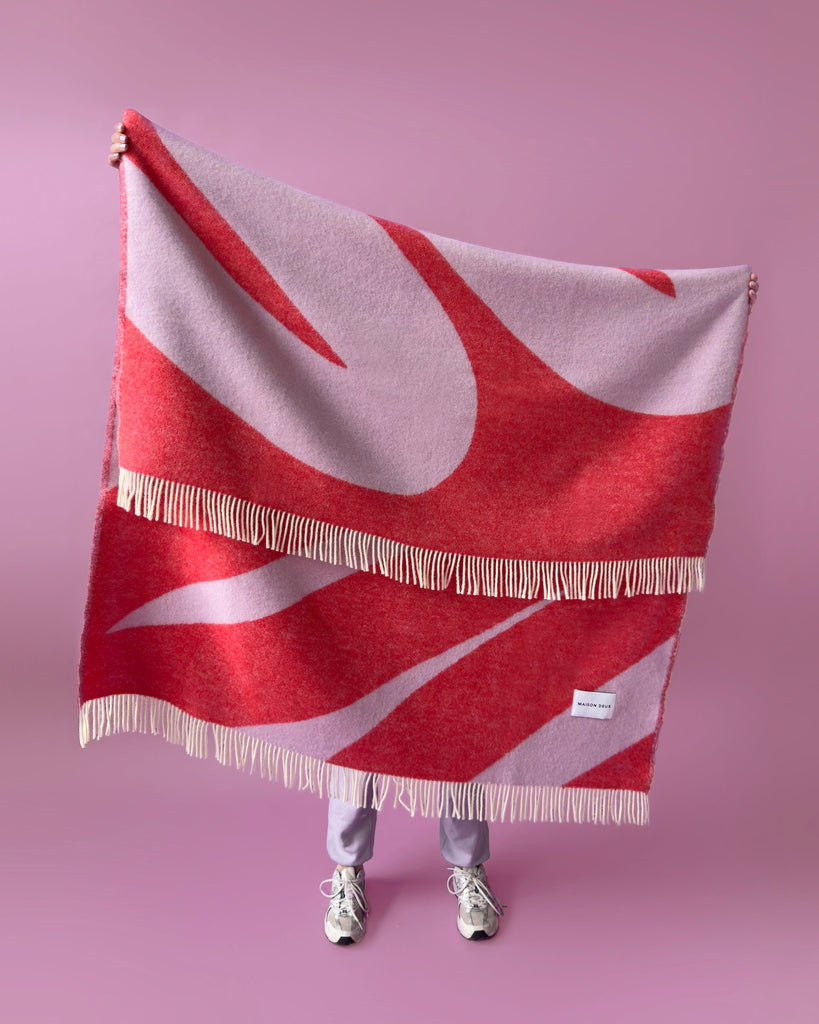 A faceless figure holding up an oversized vibrant throw with a swirling lined pattern in vibrant shades of red and purple 