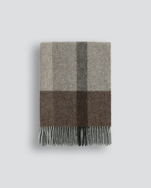 The Brunswick nz wool plaid throw blanket folded to show varied tones of grey, black and brown, and a tasseled fringe