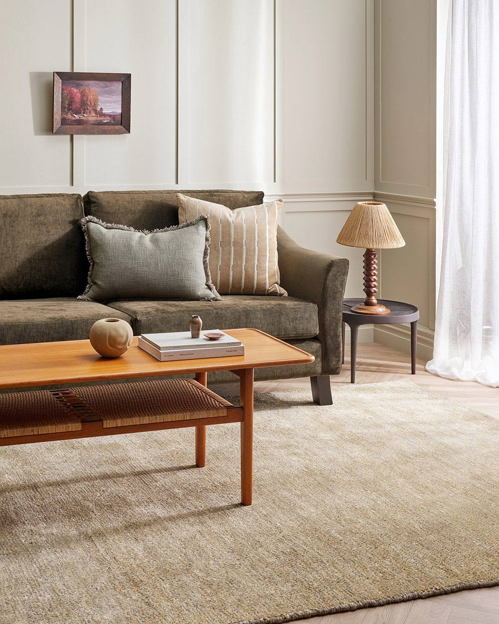 A living room with a sandy brown floor rug and a green couch. On the couch is a gorgeous beige velvet cushion with a linear striped pattern, by Baya. 