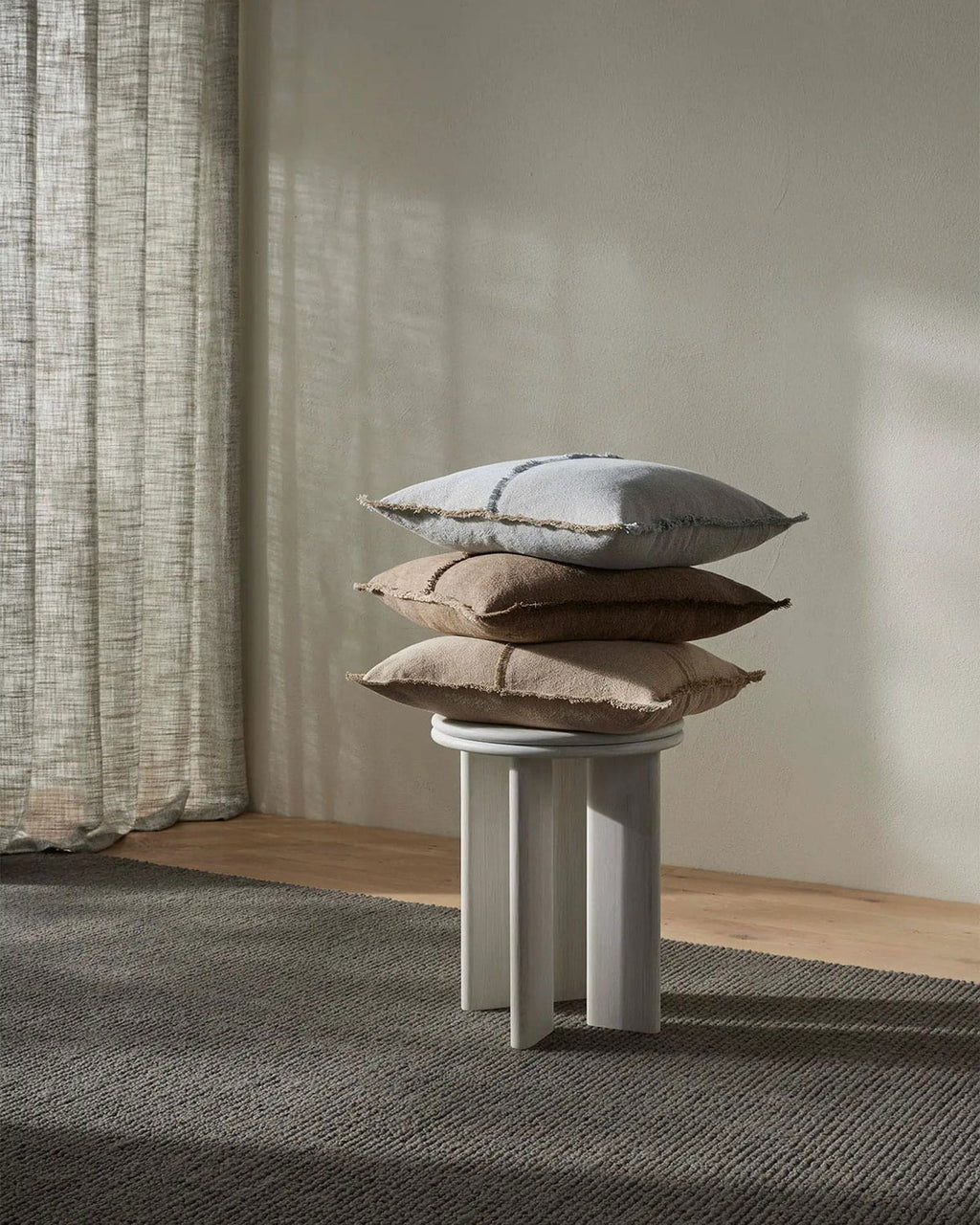 Stack of pillows on a small round table against a neutral wall with curtains.
