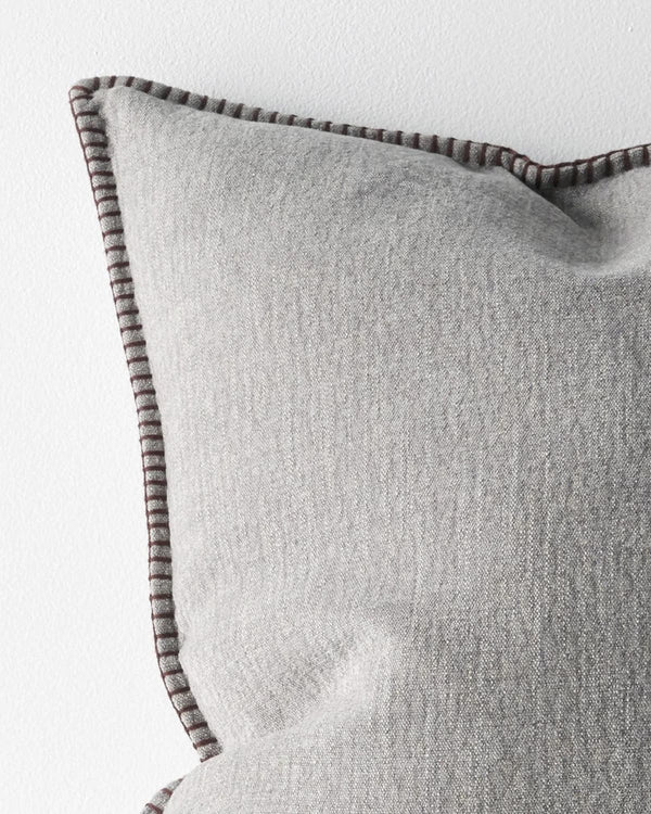 Close up corner of a grey textured cushion with a dark brown stitch detail on a light grey background