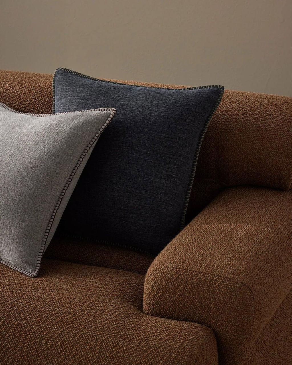 Two designer cushions in grey and blue on a brown sofa against a beige wall