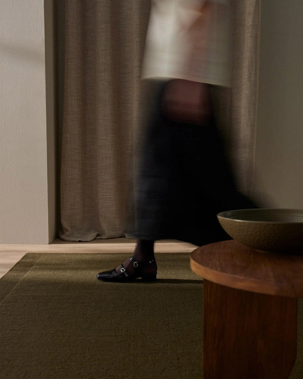 Person walking in a room with a curtain , wooden table and wool floor rug in a deep olive shade 