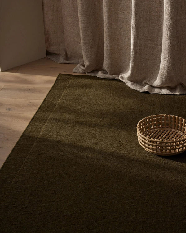 The Cassio Jasper NZ wool floor rug in a deep olive shade seen with a linen curtain