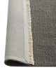 Gray rug with a textured pattern and white fringe on a plain background, the corner flipped to see the underside. 