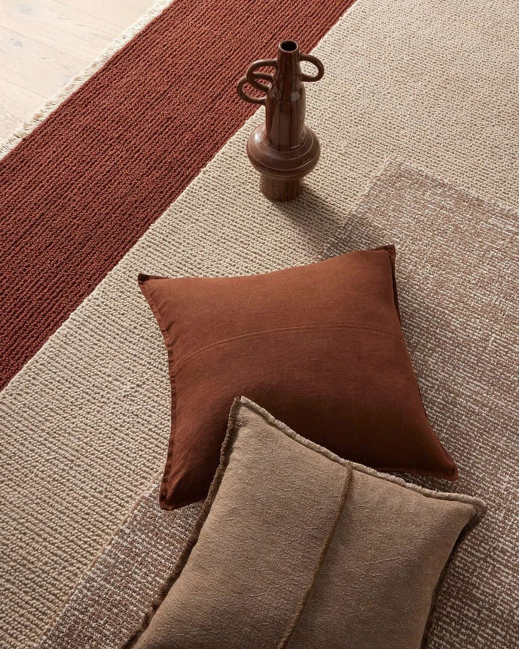 Brown and beige pillows on several warm toned floor rugs with vase. 