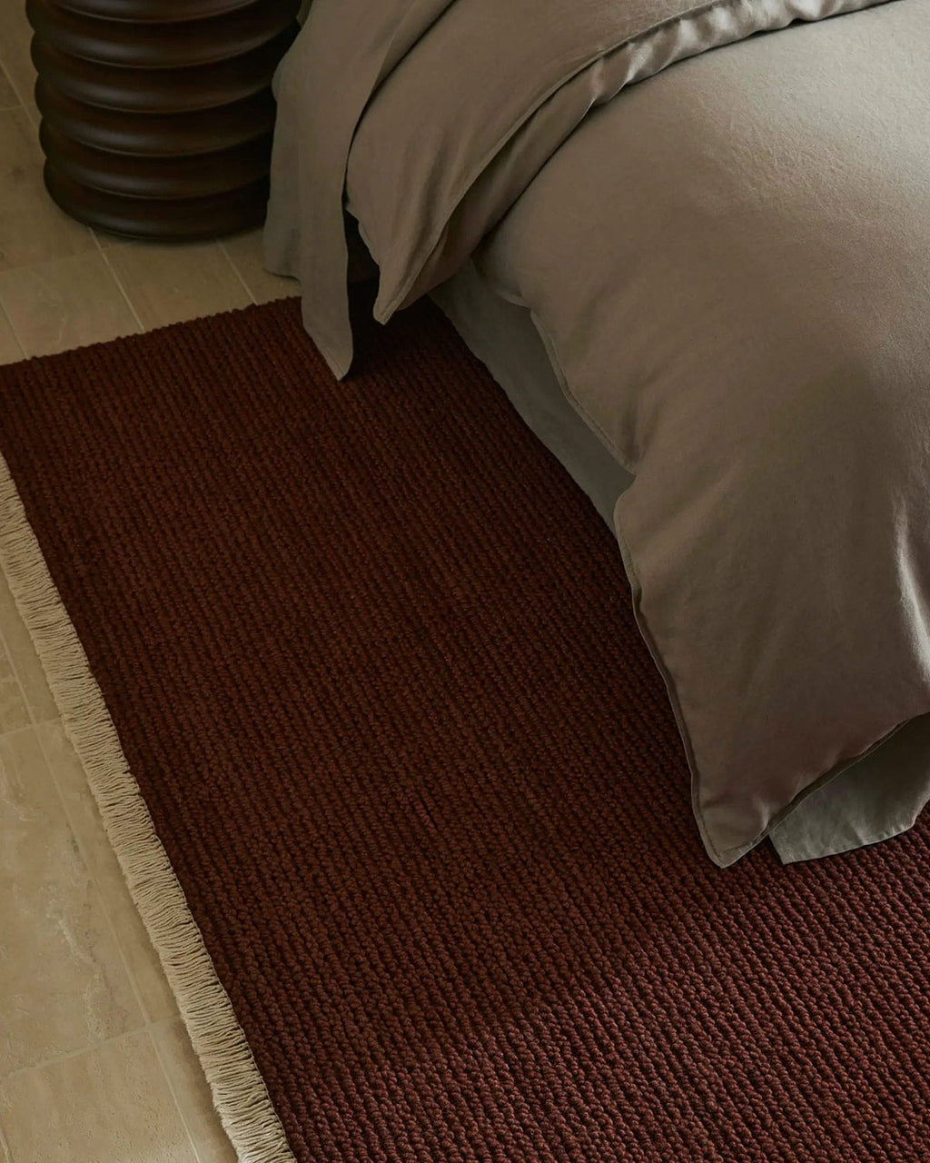 Dark red textured rug on a wooden floor next to a bed with beige bedding.