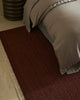 Dark red textured rug on a wooden floor next to a bed with beige bedding.