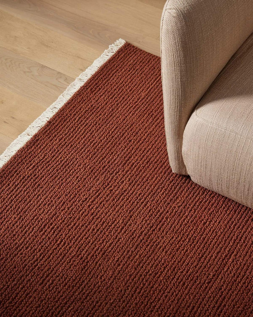 Red-brown 100% wool rug on a wooden floor with a beige sofa corner.
