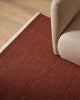Red-brown 100% wool rug on a wooden floor with a beige sofa corner.