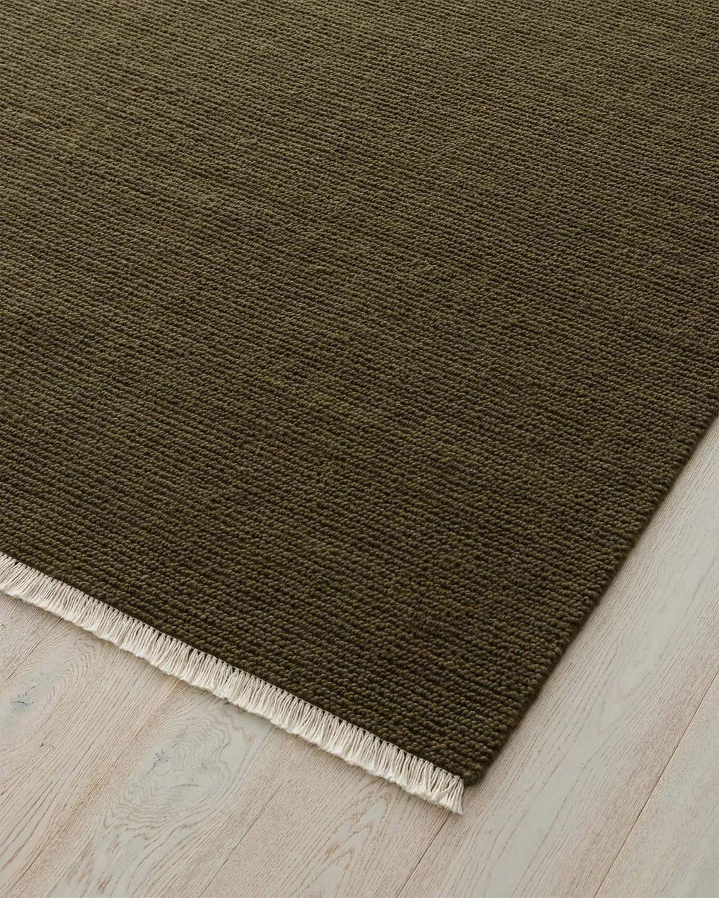 Green brown textured rug with white fringe on a light wood floor