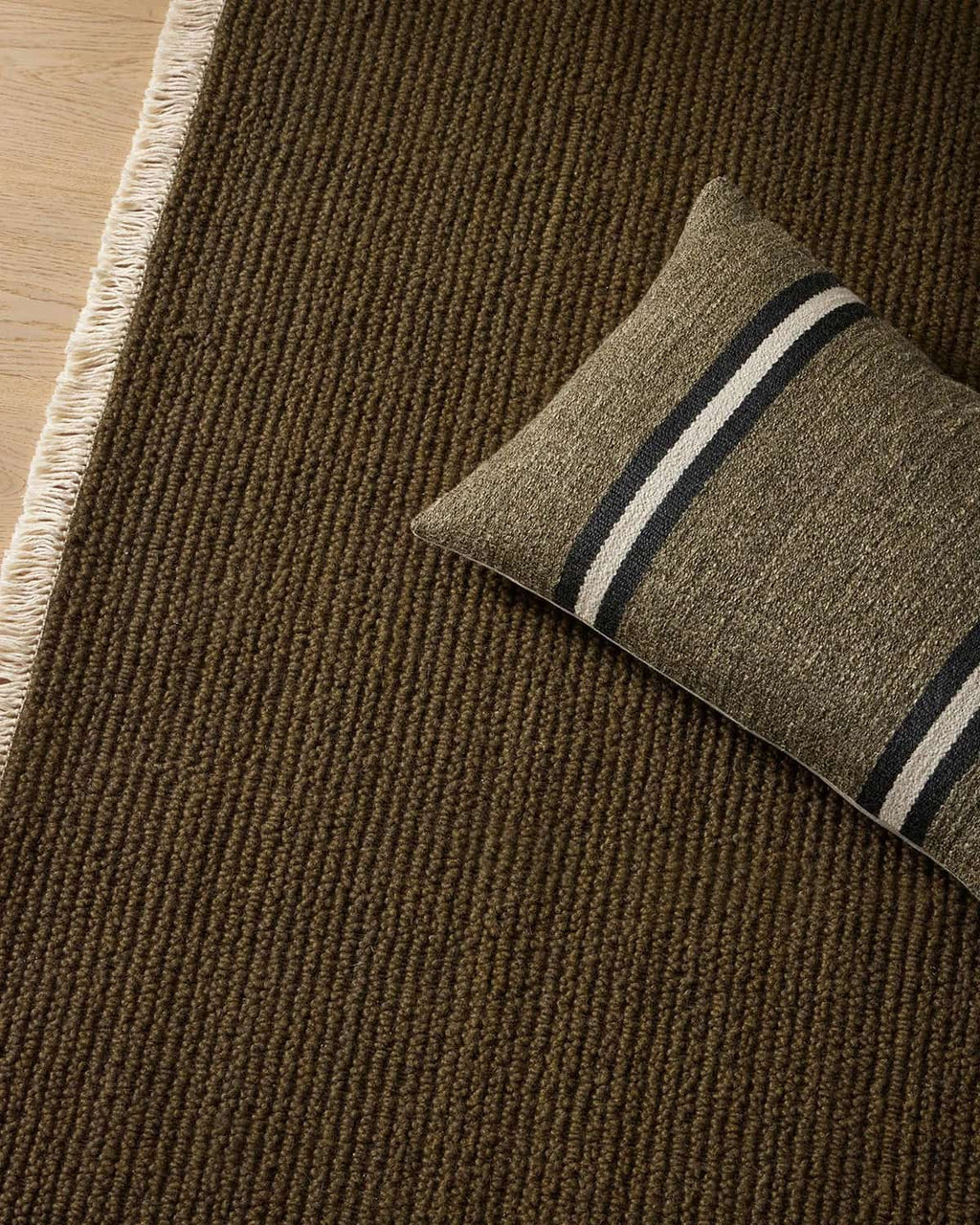 Green brown 100% wool textured rug with a pillow featuring stripes on a wooden floor.