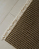 Brown textured rug with white fringe on a light wooden floor