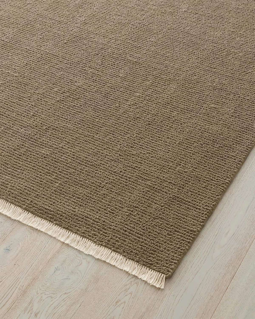 Brown textured rug with a fringed edge on a light wood floor.