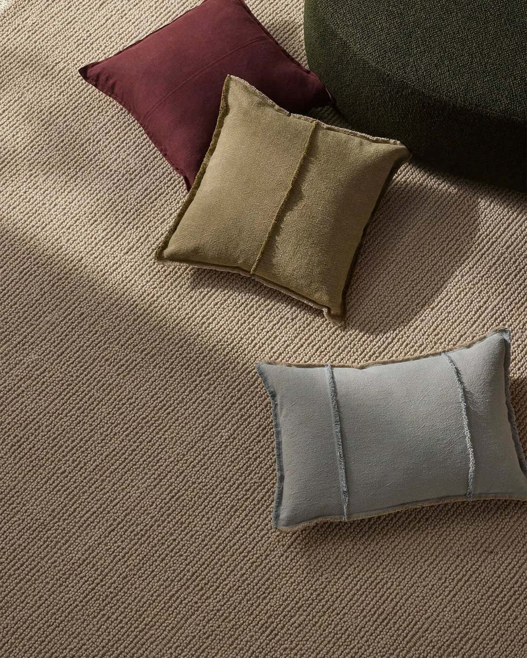 Three cushions in maroon, green, and gray on a textured neutral beige floor rug