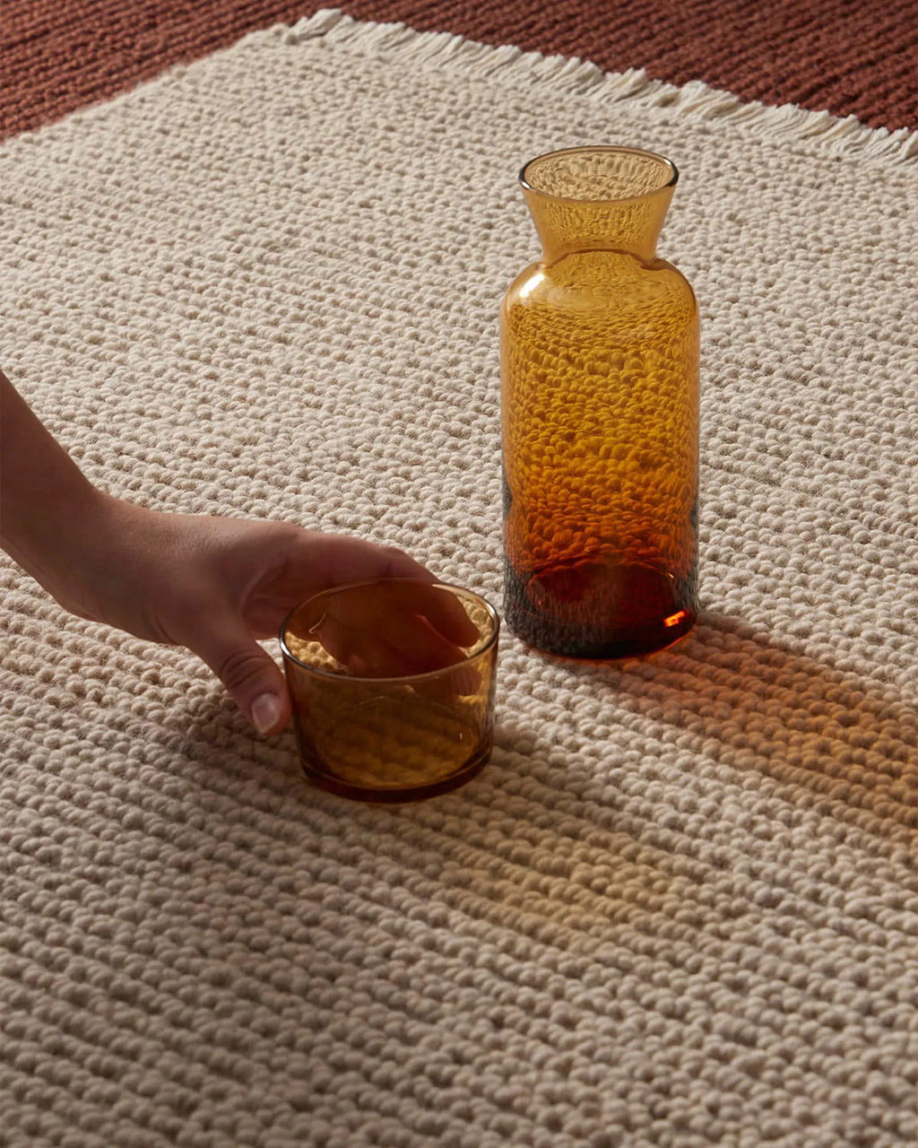 Two amber-colored glasses on a textured beige wool floor rug with a hand reaching towards one of them.