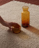 Two amber-colored glasses on a textured beige wool floor rug with a hand reaching towards one of them.