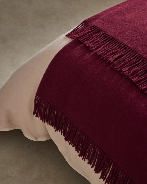 Weave Home wool throw blanket in colour 'rhubarb' - a rich deep red, shown on the end of a bed