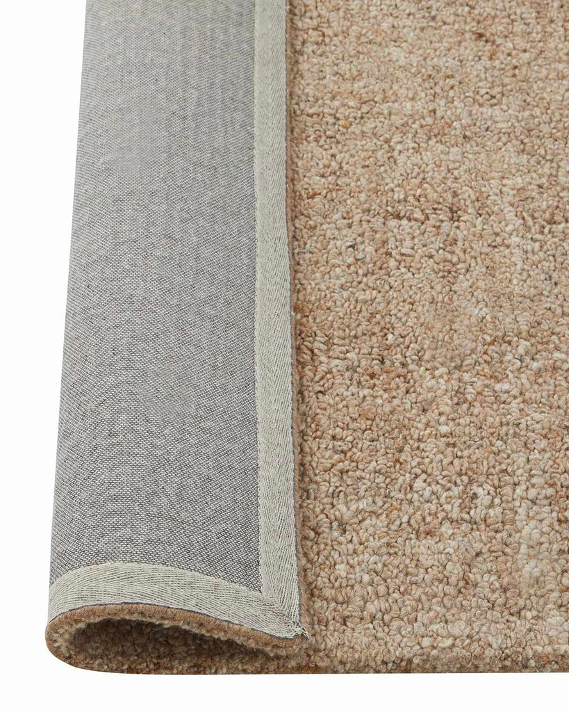 A close up image of a warm beige loop pile floor rug with the edge flipped over to show the durable cotton backing. 