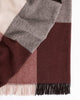 A folded throw blanket in 100% wool with a checked pattern in shades of warm chocolate brown, soft pink and berry tones, with twisted fringing. 