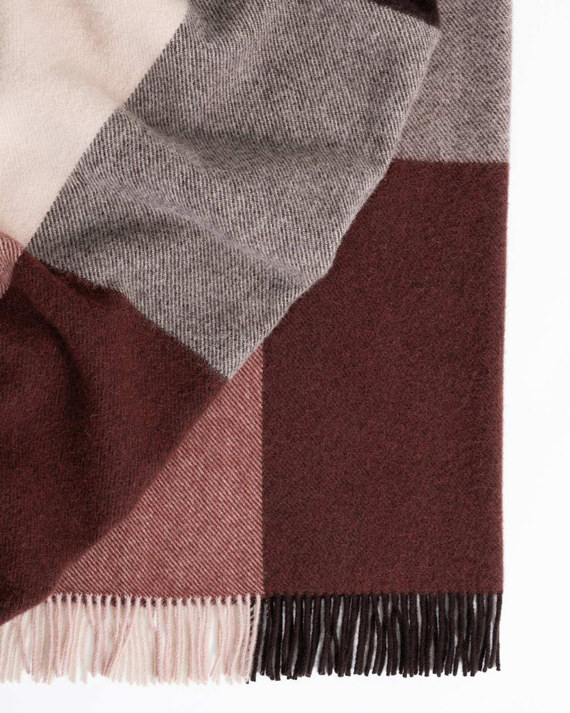 A folded throw blanket in 100% wool with a checked pattern in shades of warm chocolate brown, soft pink and berry tones, with twisted fringing. 