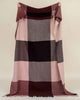 A large throw blanket of 100% wool in berry tones with a big check pattern and fringed edges, held up so the full throw is visible. 