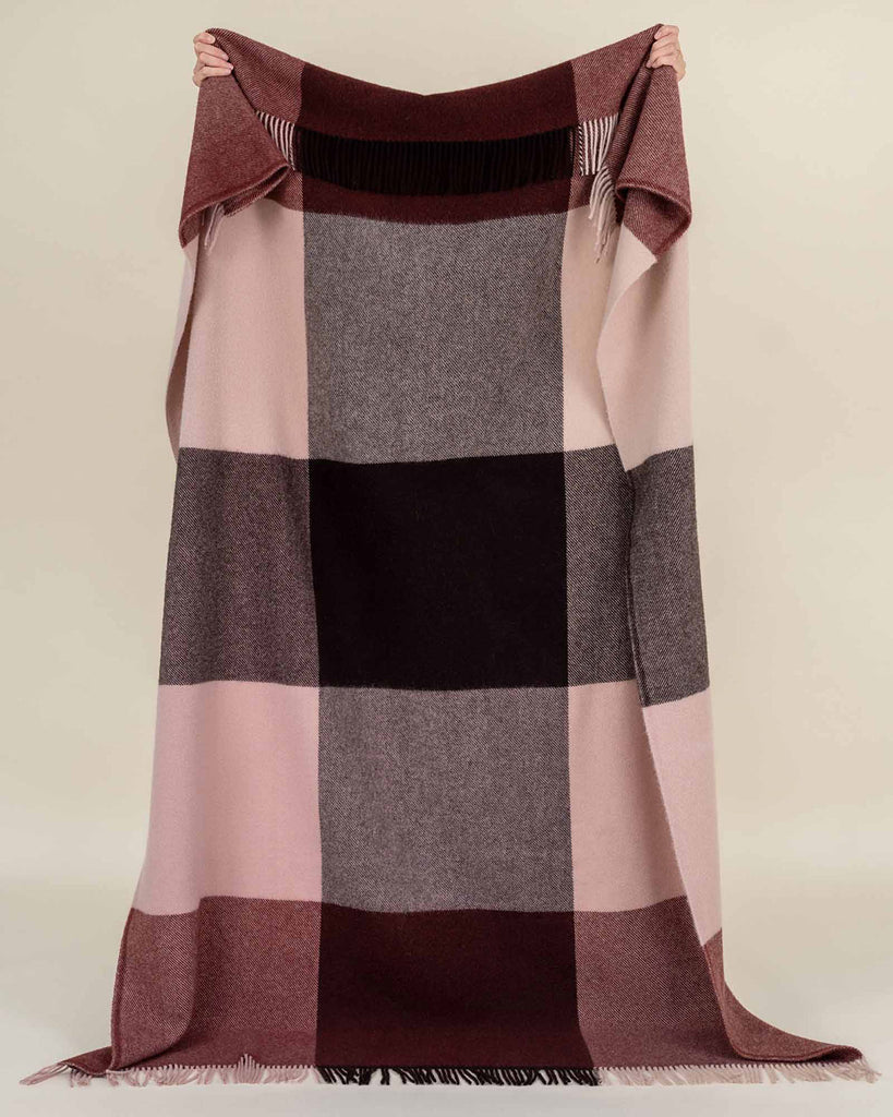 A large throw blanket of 100% wool in berry tones with a big check pattern and fringed edges, held up so the full throw is visible. 