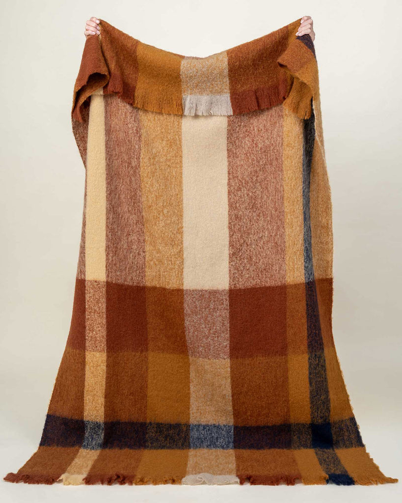 A large throw blanket held up to see the full image, in checked cream, orange, and brown, with fringe edges. 
