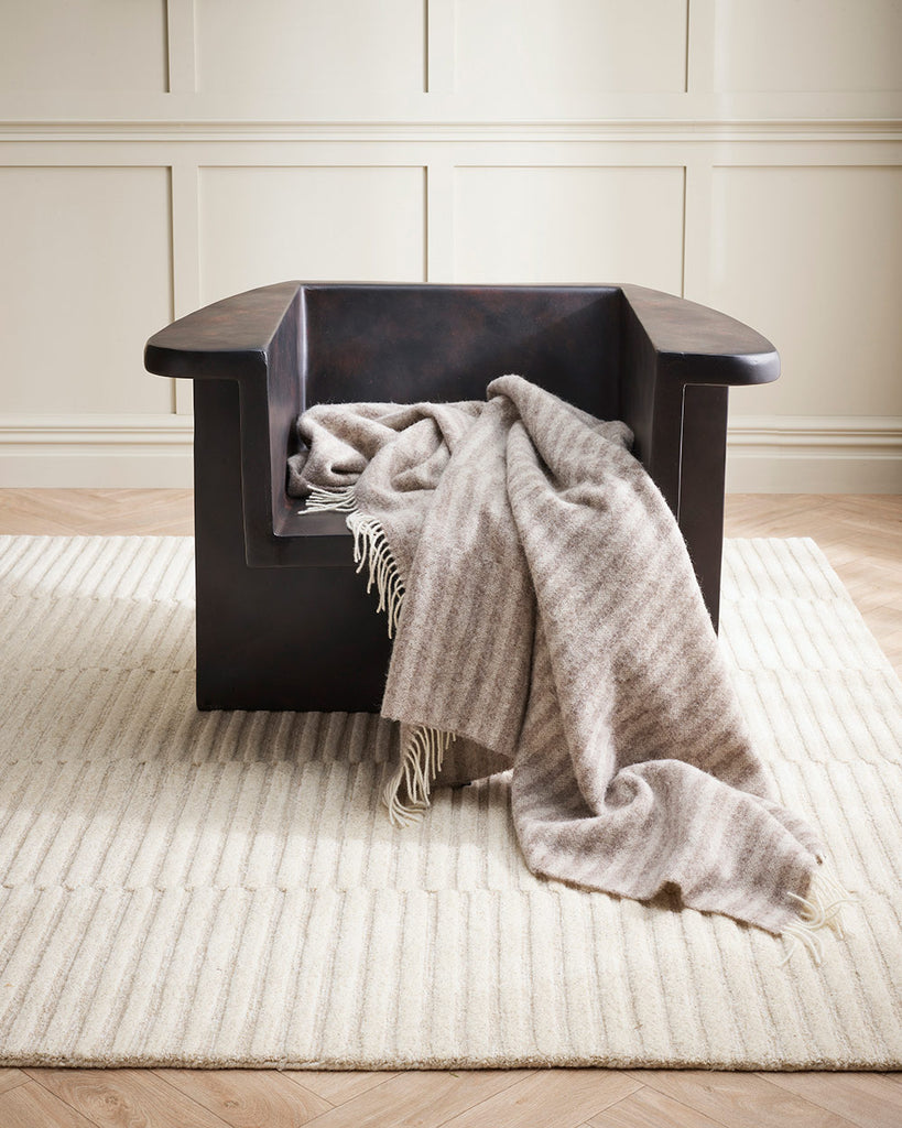 A modern style chair in dark wood with a neutral striped throw blanket, atop a stunning neutral cream toned cut and loop pile linear patterned floor rug. 