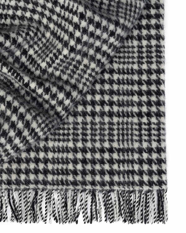 A folded throw blanket in 100% wool with a sophisticated black and white houndstooth check pattern and twisted fringe edging.