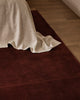 Baya Bastion NZ wool floor rug in Claret, close view at the foot of a bed with a linen throw, on a timber floor.