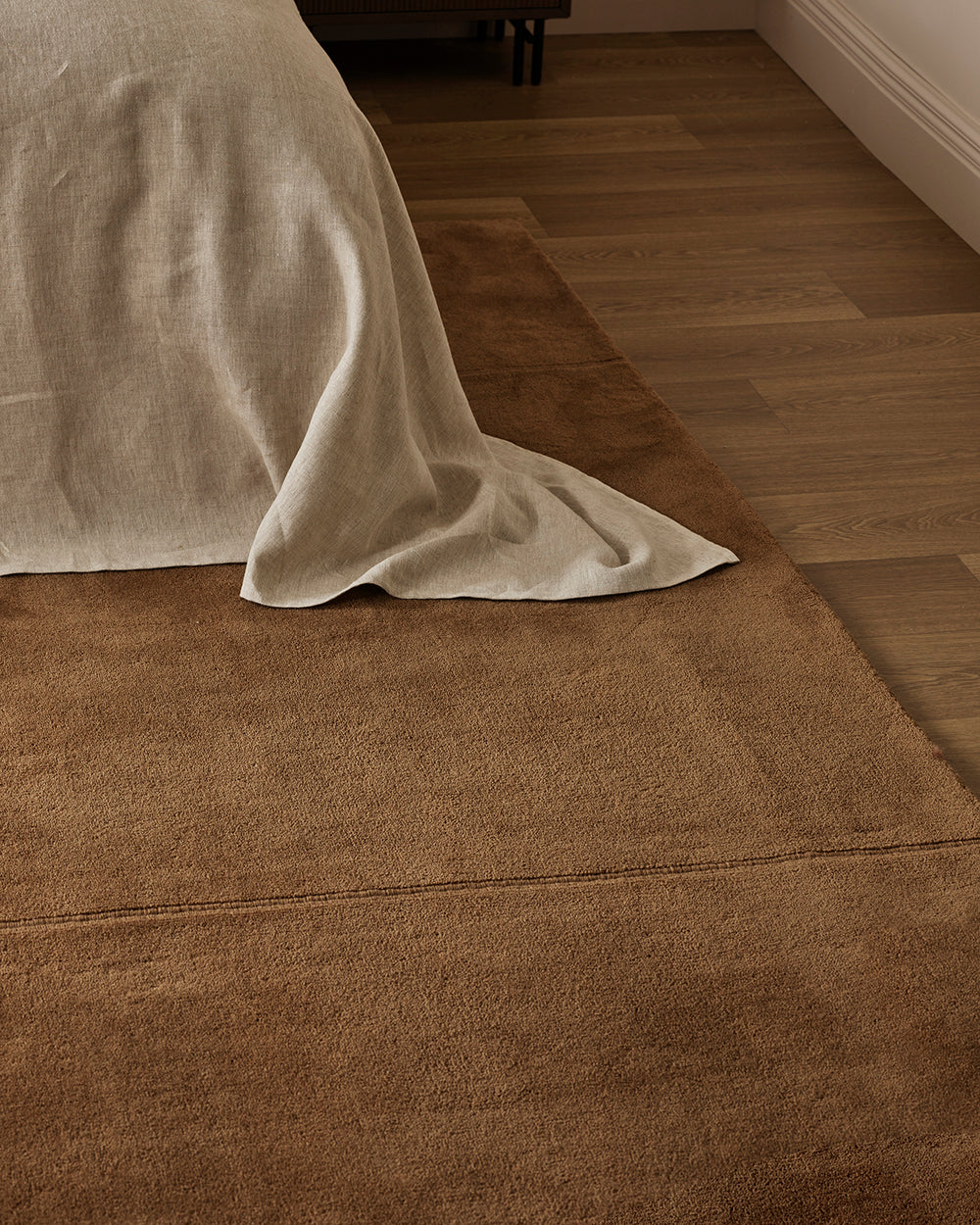 Baya Bastion NZ wool floor rug in Hazelnut, close view at the foot of a bed with a linen throw, on a timber floor.