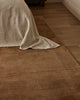 Baya Bastion NZ wool floor rug in Hazelnut, close view at the foot of a bed with a linen throw, on a timber floor.