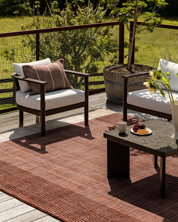 Baya Bedrock Indoor and Outdoor Floor Rug in Claret, styled on an outdoor deck under a timber armchair with a striped cushion, beside a potted plant and a low stone-topped coffee table set with fruit and sunflowers.