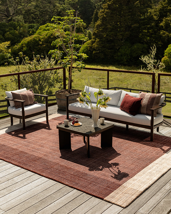 Baya Bedrock Indoor and Outdoor Floor Rug in Claret, full-width view on a deck beneath a sofa, armchair and coffee table, showing the rug's rust-red field and pale woven border.
