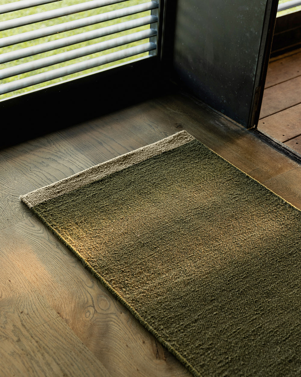 Baya Carla NZ wool floor mat in Military, styled at an entrance beside a window in afternoon light.