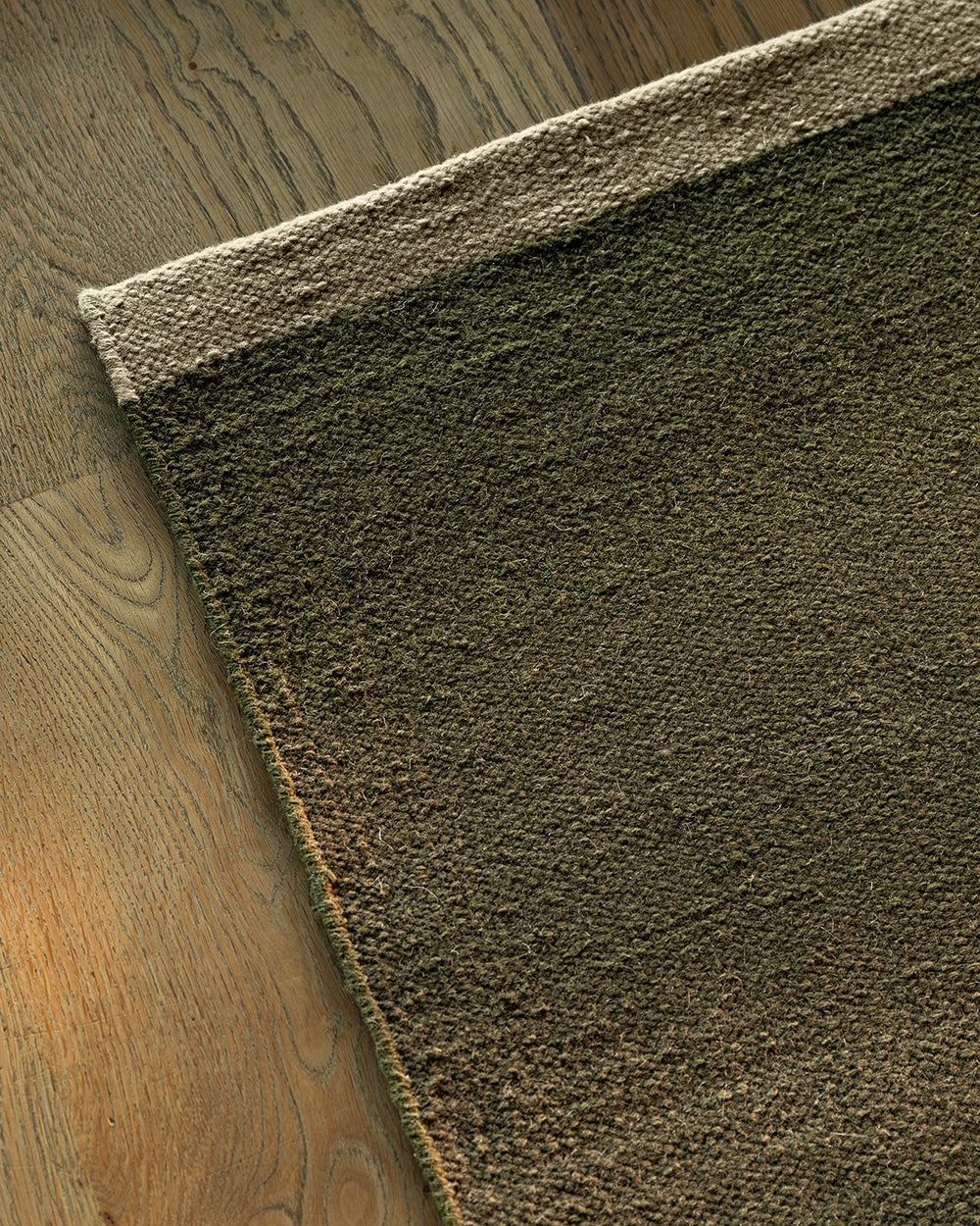Baya Carla NZ wool floor mat in Military, folded corner showing the weave and underside on a wood floor.