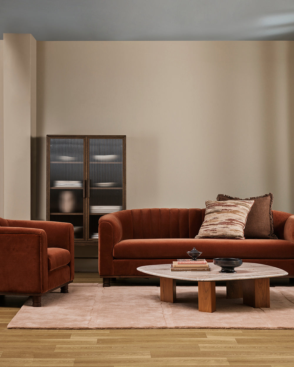 Wider view of the Baya Dover Chocolate cushion styled on a rust velvet sofa and matching armchair