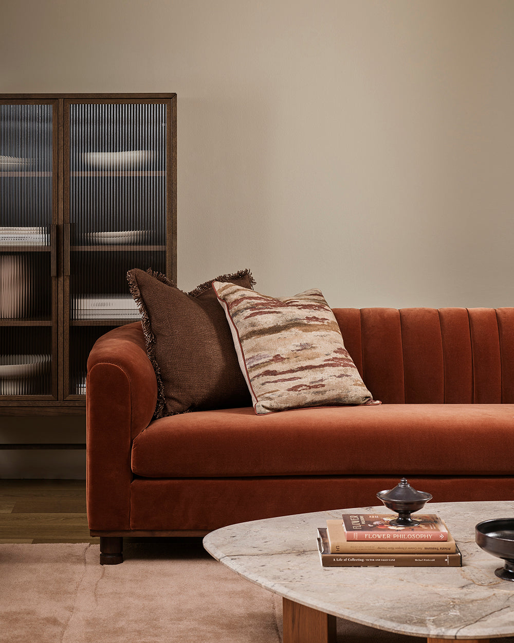 Baya Dover Chocolate cushion styled on a rust velvet sofa with a patterned cushion, in front of a ribbed glass cabinet