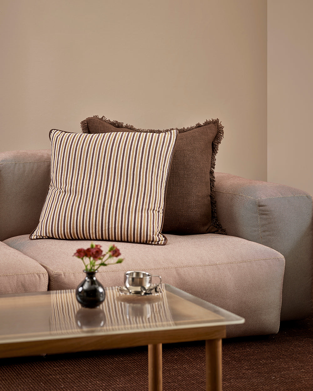 Baya Dover Chocolate cushion styled on a grey sofa alongside a striped cushion, close view with a vase of flowers on the coffee table
