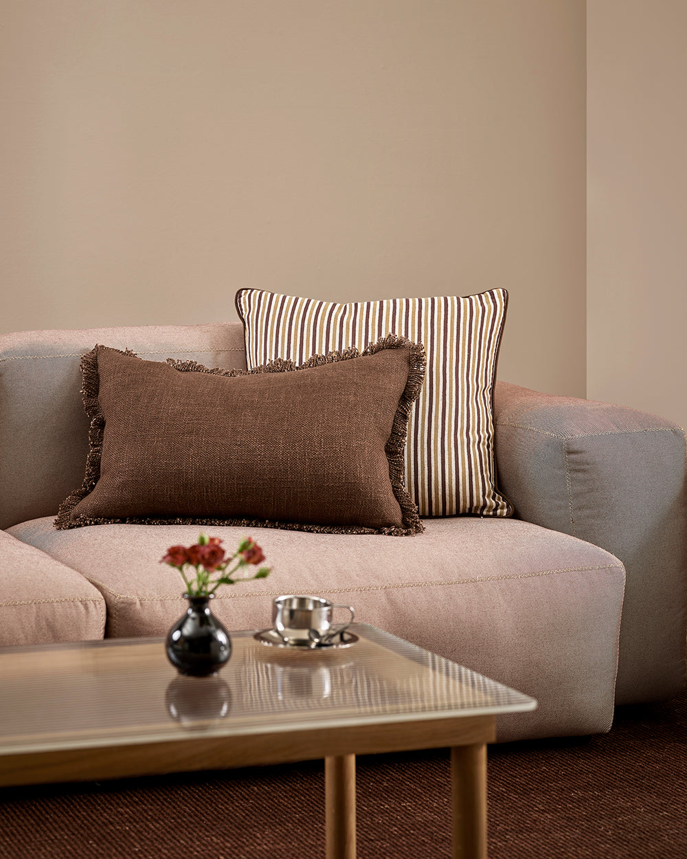 Baya Dover Chocolate lumbar cushion styled on a grey sofa alongside a striped cushion, close view with a vase of flowers on the coffee table