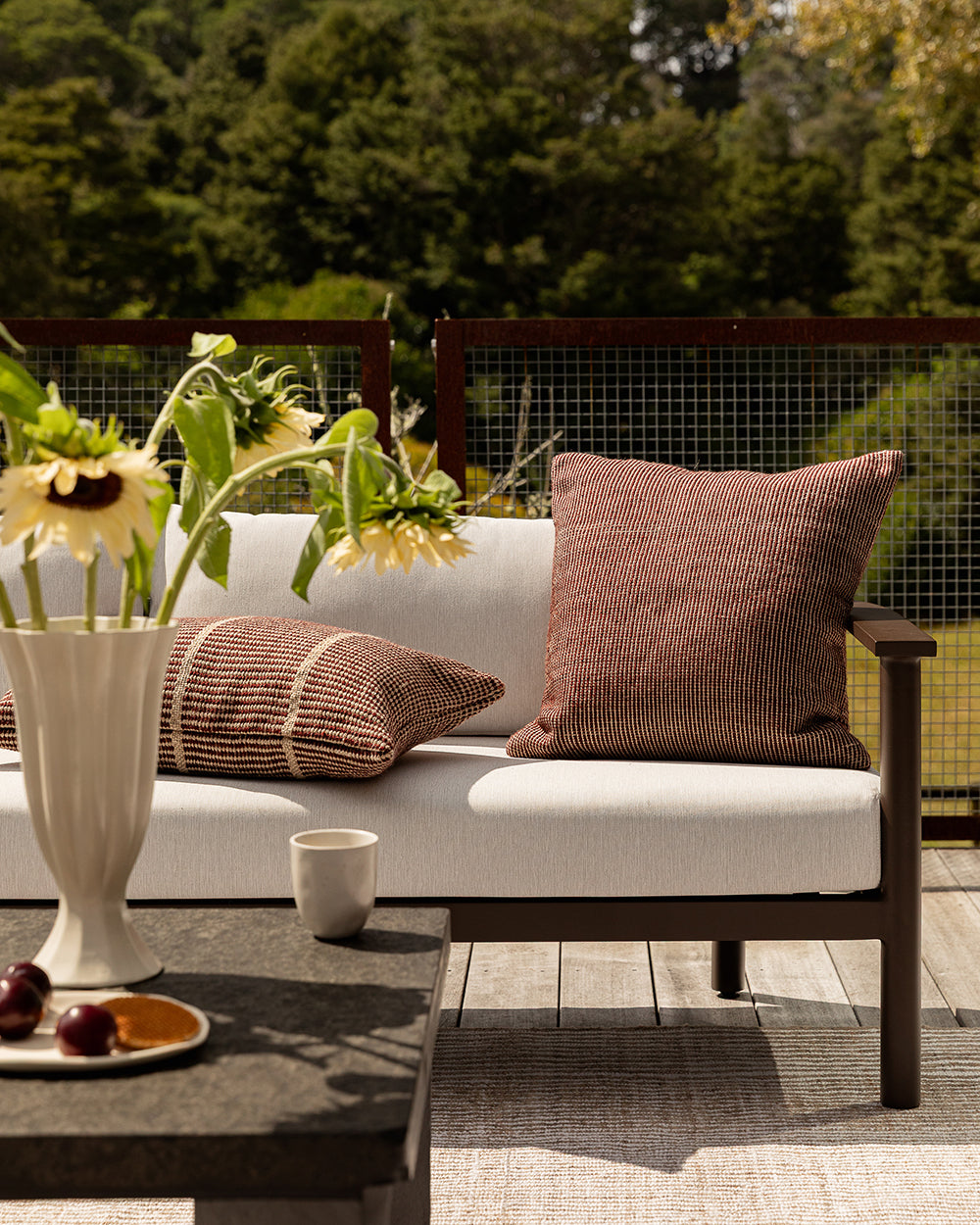 Baya Dune outdoor cushion in Claret styled on an outdoor sofa with a striped lumbar cushion, sunflowers and fruit on the side table