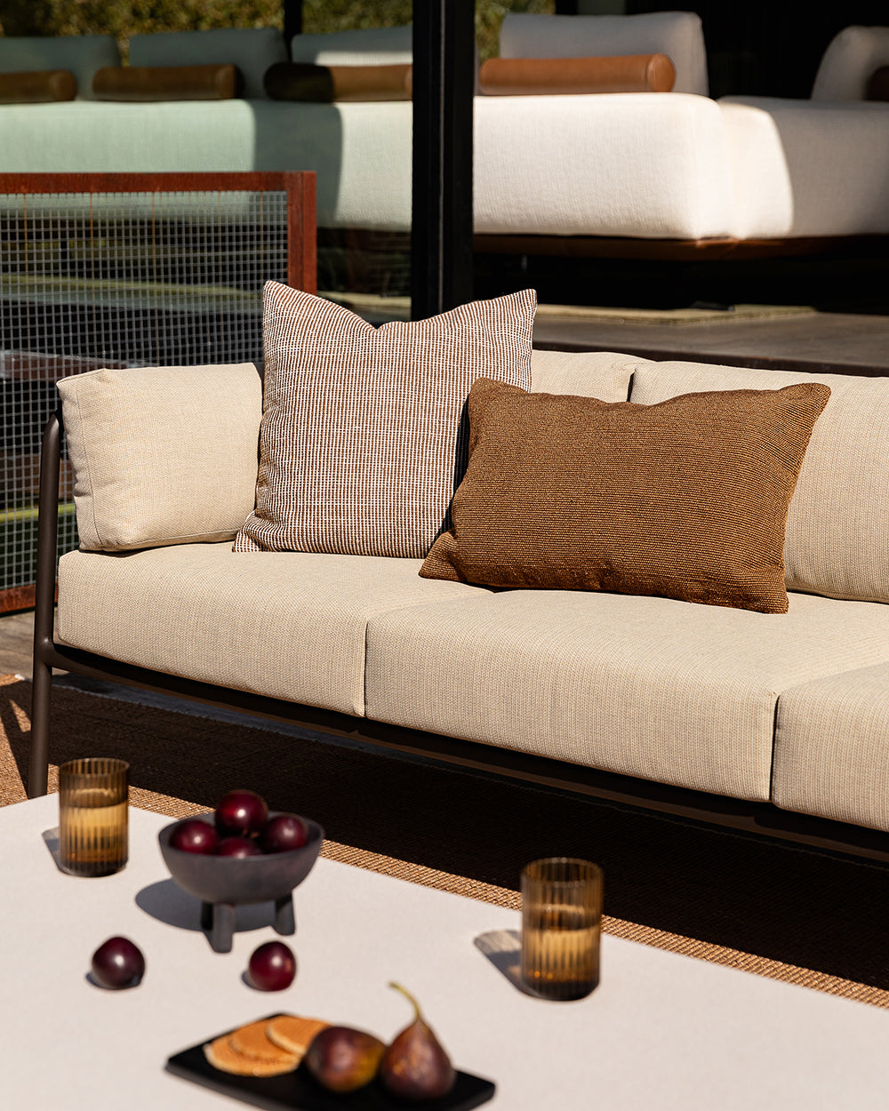 Baya Dune outdoor cushion in Tobacco styled on an outdoor sofa beside a solid rust-brown lumbar cushion, with a styled coffee table setting