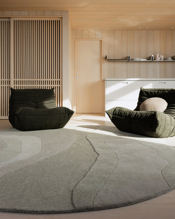 Baya Edgewater NZ wool floor rug in Pistachio, round, styled in a timber-panelled room with two dark green lounge chairs.