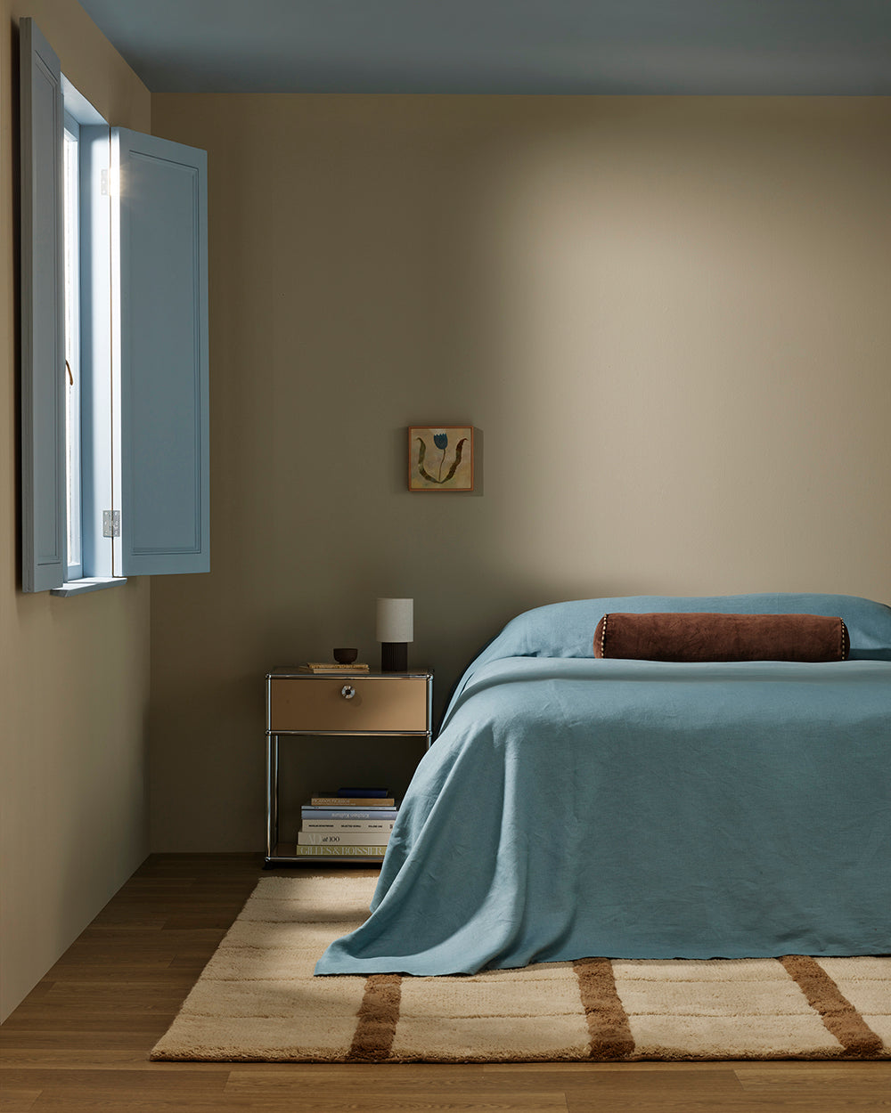 Baya Elliot cotton velvet bolster cushion in Chocolate, on a bed in a bedroom with an open window