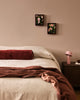 Baya Elliot cotton velvet bolster cushion in Oxblood, on a bed styled with a brown throw and framed botanical art